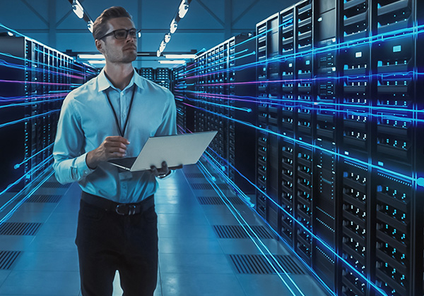 A male employee holding his laptop looking a row of datacenter.