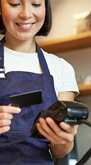 Young female worker in apron about to swipe credit card through a payment processor.