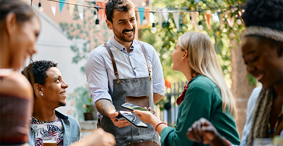 A man wearing an apron holding out his phone to a woman, while their colleagues looks on.